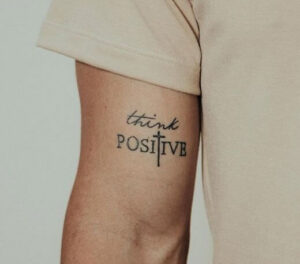 tatuaggio uomo sul bicipite con scritto think positive con croce